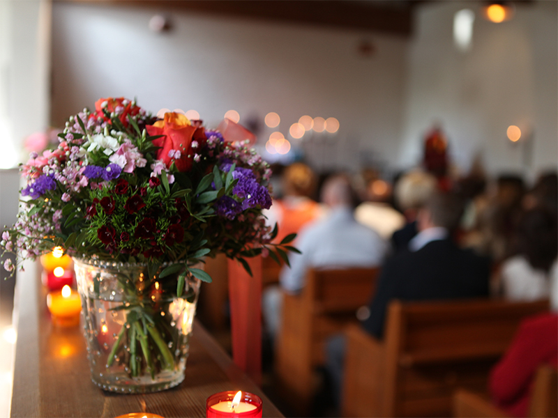Ein bunter Blumenstrauß in einer Glasvase und brennende Kerzen stehen im scharfen Fokus auf einem Holzsims. Im unscharfen Hintergrund sind Trauergäste in den Bänken einer hellen Kapelle zu sehen.