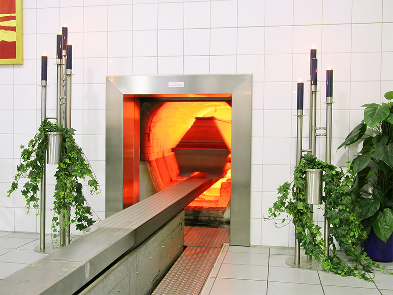 Ein Sarg wird im Krematorium zur Einäscherung in den hell beleuchteten Ofen eingefahren, eingerahmt von dekorativen Kerzenständern und Efeu.