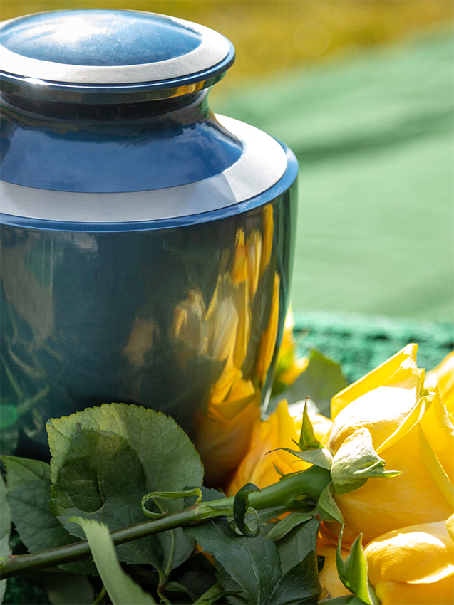 Eine glänzend dunkelblaue Bestattungsurne mit silbernem Rand, vor der gelbe Rosen als Symbol des Gedenkens liegen.