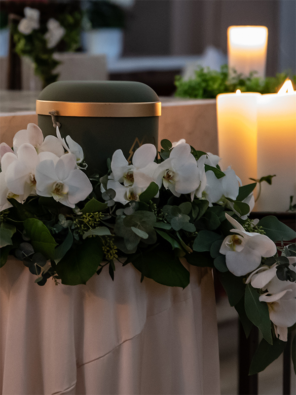Eine matte, dunkelgrüne Urne mit Goldakzent, eingebettet in ein Gesteck aus weißen Orchideen und grünen Blättern neben brennenden Kerzen.