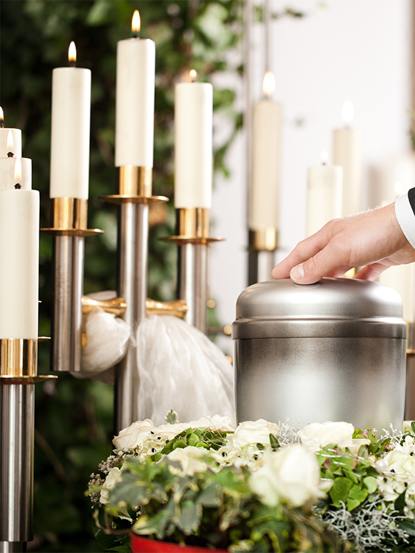 Detailansicht einer Hand, die sanft auf einer silbernen Urne ruht, umgeben von weißen Blumen und festlichen Kerzenleuchtern.