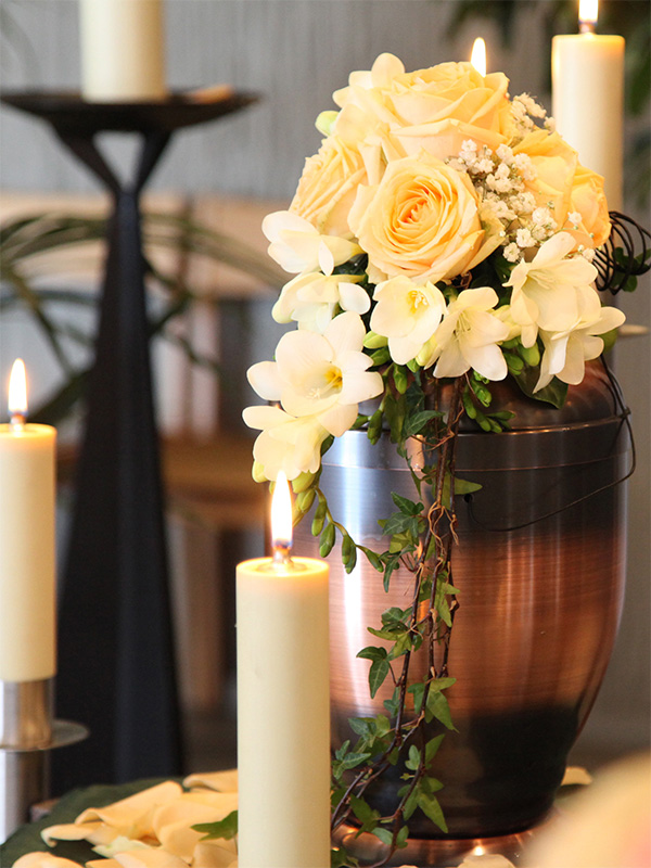 Eine kupferfarbene Metallurne, geschmückt mit einem fließenden Gesteck aus gelben Rosen und Efeu im warmen Licht von Stumpenkerzen.